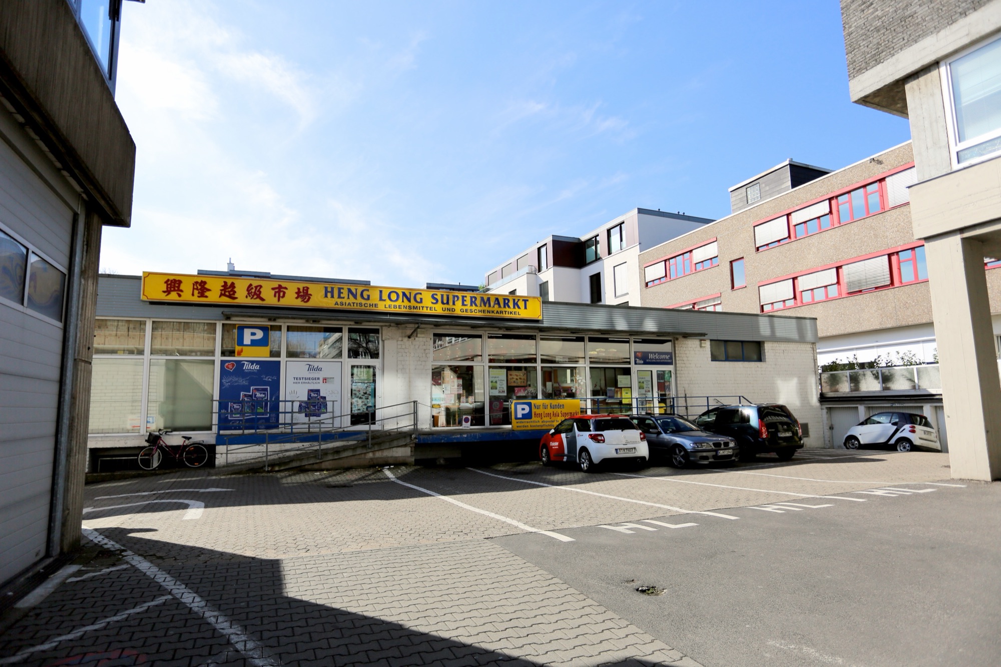 Außenansicht des Heng Long Supermarkts mit gelbem Schild „興隆超級市場 – Heng Long Supermarkt“ und Parkplätzen davor